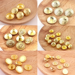 May include: Assortment of gold-toned metal buttons with various designs. Some buttons have a raised design, while others have a flat surface. The buttons are arranged in a grid pattern on a wooden surface.