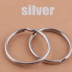May include: Two silver key rings, one slightly larger than the other, are shown on a brown surface. The word "silver" is printed in white above the key rings.