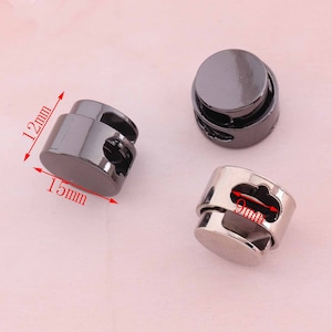 Pode incluir: Três travas de cordão de metal preto com um diâmetro de 12 mm e uma altura de 15 mm. As travas têm uma forma redonda e um furo no centro para enfiar o cordão.