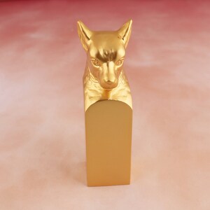 May include: A gold-coloured animal head sculpture with a rectangular base. The sculpture is of a dog or wolf with a serious expression.