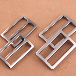 May include: Two silver metal rectangular belt buckle slides. The slides are stacked on top of each other.