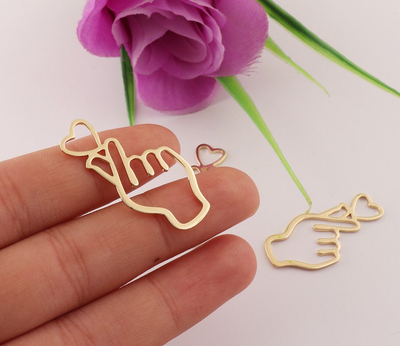 Op de afbeelding: Twee gouden metalen bedels in de vorm van een hand die een pinkbelofte doet met een hart. De bedels zijn perfect voor het maken van sieraden of knutselen.