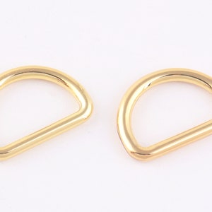 Puede incluir: Dos anillos en D de metal de color dorado. Los anillos son lisos y brillantes.