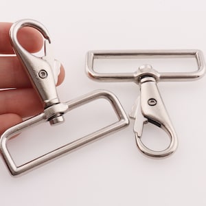 Könnte beinhalten: Zwei silberne Metall-Schwenkhaken mit rechteckigen Schlaufen. Die Haken sind zum Befestigen an Gurten, Gürteln oder anderen Gegenständen gedacht.