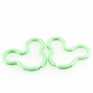 Puede incluir: Dos llaveros verde claro con forma de cabeza de Mickey Mouse. Los llaveros son de metal y tienen un diseño de doble bucle. Los llaveros están sobre un fondo blanco.