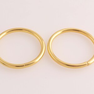 Puede incluir: Dos aros de metal de color dorado, cada uno con un diseño circular sin costuras. Los aros son ligeramente diferentes en tamaño, con el aro más grande a la derecha y el más pequeño a la izquierda.