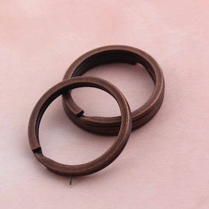 Può includere: Due portachiavi in ​​rame antico, uno leggermente più grande dell'altro, sono impilati l'uno sull'altro. Gli anelli sono lisci e hanno una finitura leggermente usurata.