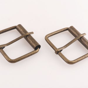 May include: Two antique gold-toned metal belt buckles. Each buckle has a square frame with a rounded, slightly curved, metal pin.