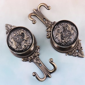 Könnte beinhalten: Zwei antike goldfarbene Metallhaken mit verzierten Designs. Jeder Haken verfügt über eine runde Platte mit einem geprägten Drachenmotiv. Die Haken eignen sich perfekt zum Aufhängen von Vorhängen oder anderen Dekorationsartikeln.