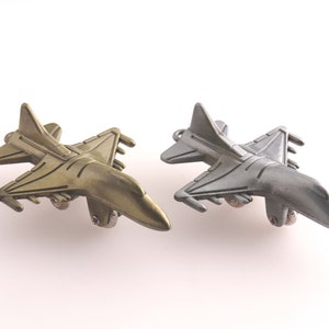 Puede incluir: Dos aviones de combate metálicos en miniatura, uno dorado y otro plateado, están colocados uno al lado del otro sobre un fondo blanco.
