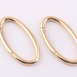 Puede incluir: Dos cierres metálicos ovalados de color dorado. Los cierres están abiertos y listos para ser unidos a una bolsa u otro artículo.