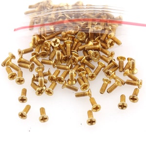 May include: A large quantity of small, gold-coloured screws. The screws have a Phillips head and are scattered on a white surface.