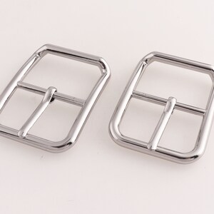 May include: Two silver metal belt buckles. The buckles are rectangular with rounded corners and have a smooth, polished finish.