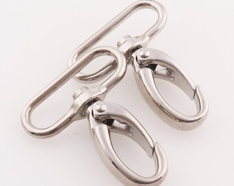 Op de afbeelding: Twee zilveren metalen draaibare gespen met een rechthoekige lus en een D-ring. De gespen worden vaak gebruikt om banden of kettingen aan tassen, handtassen of andere accessoires te bevestigen.
