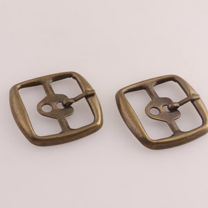 May include: Two antique brass-colored square buckles. Each buckle has a central design with two eyelets and a hinged clasp. The buckles are likely for belts or other accessories.