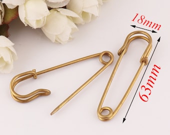 May include: Two antique brass safety pins. One pin is open, the other is closed. The closed pin measures 63mm long and 18mm wide. The pins are on a white surface with white flowers in the background.