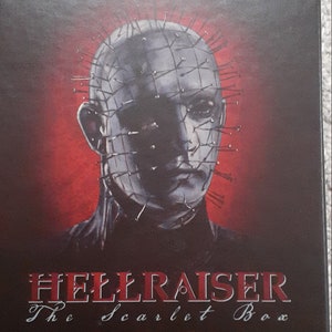 May include: A black box set for the Hellraiser horror film franchise. The box features a red background with a close-up image of Pinhead, the main antagonist, with his signature hooked chain and pin-covered head. The text "The Scarlet Box" is printed in white on the box, along with the numbers "I II III" to indicate the included films.