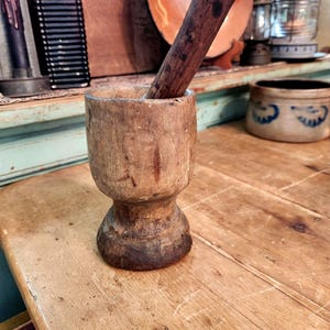 Könnte beinhalten: Ein verwittertes Mörser- und Stößel-Set aus Holz, bestehend aus einer zylindrischen Schale mit breitem Boden und einem passenden dunkelbraunen Holzstößel, der darin liegt. Das Holz zeigt eine natürliche Maserung und Gebrauchsspuren.