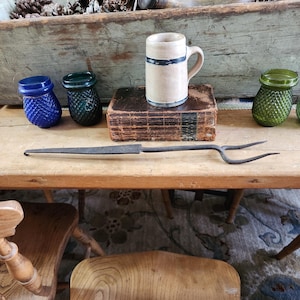 May include: A rustic still life featuring a collection of vintage items. A tan mug with a blue stripe sits atop a worn book. Beside it are blue and green glass jars, and a long, wrought-iron fork. The items are arranged on a wooden table.
