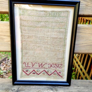 May include: Framed antique textile art with a black frame. The textile features embroidered letters and a decorative border in red and green on a beige background. The piece is displayed on a wooden surface.