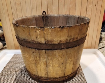 Puede incluir: Un cubo de madera envejecida con aros metálicos. El cubo tiene forma cilíndrica con la parte superior ligeramente ensanchada. La madera es de color marrón claro con vetas visibles y algunas manchas oscuras. Un asa metálica está fijada al borde.