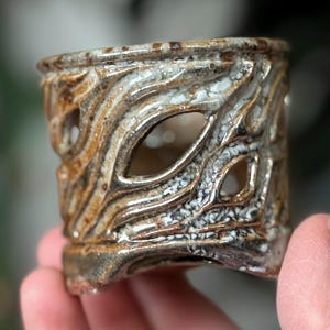 May include: A handcrafted ceramic candle holder with a unique, textured design. The holder features a brown and white color scheme with cut-out patterns. The intricate details and earthy tones create a rustic aesthetic, suitable for home decor.