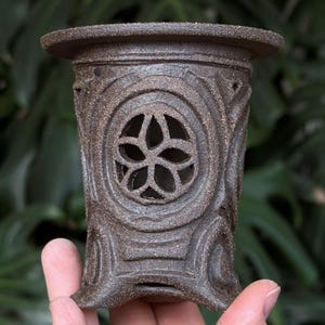 Peut inclure: Un photophore en céramique marron avec une découpe en forme de fleur. Le support cylindrique a une surface texturée et des sculptures décoratives. Le support est tenu dans une main sur fond de feuilles vertes.
