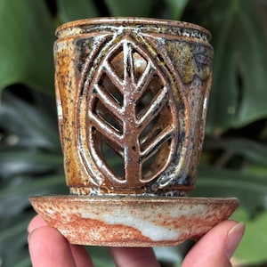May include: A handcrafted ceramic candle holder featuring a leaf-shaped cut-out. The cylindrical holder sits on a matching saucer. The colors are earthy browns, tans, and white, with a glazed finish. A decorative item.