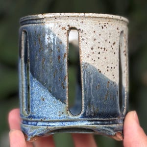 May include: A handmade ceramic candle holder with a cylindrical shape. The holder features a two-tone design, with a speckled white top and a blue-glazed bottom. Vertical cutouts create a decorative pattern. The holder is held by a person's hand.