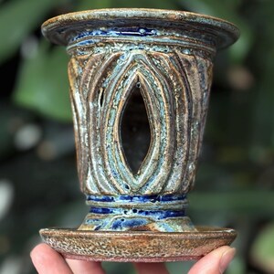 May include: A handcrafted ceramic candle holder with a unique design. The holder features a brown and blue colour scheme with an open, oval-shaped centre. The base is a small, round dish. The background is a blurred green.
