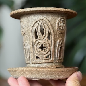 May include: A tan ceramic candle holder with a detailed gothic design. The holder features arched cutouts and cross motifs, with a circular base and top. The design is reminiscent of cathedral architecture.