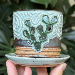May include: A light blue ceramic planter with a green leafy design and a matching saucer. The planter has a textured surface and a brown rim.