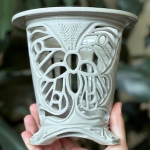 以下が含まれることがあります： 蝶のデザインが施された灰色のセラミック製のプランター。プランターは幅広の縁と4本の脚のある台座があります。