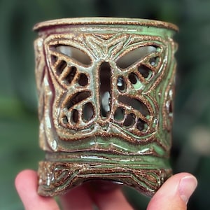 Może przedstawiać: Ręcznie robiony ceramiczny świecznik z motywem motyla. Cylindryczny uchwyt ma zielono-brązową glazurę. Motyl jest wycięty, pozwalając na prześwit światła. Podstawa ma dekoracyjny wzór.