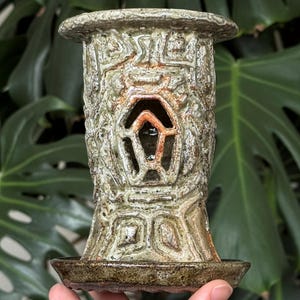 May include: A ceramic candle holder with a textured, carved design in shades of green and brown. The holder has a cylindrical shape with a flared top and a small, round base. It features a geometric cutout design.