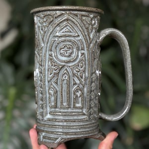 May include: A gray ceramic mug with a handle, featuring an embossed Gothic design. The mug has a rectangular base and a detailed pattern of arches, circles, and floral elements. The mug is held in front of a blurred green background.
