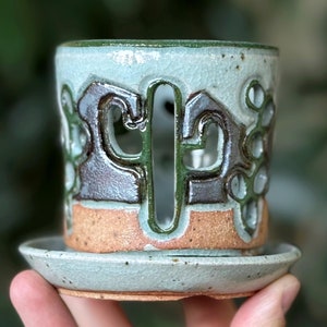 May include: A light blue ceramic planter with a cutout design of a cactus. The planter has a brown and green glaze and a matching saucer.