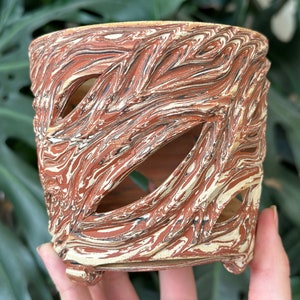 Puede incluir: Un pequeño macetero de cerámica rojo y blanco con un diseño único de mármol y tres pequeños pies. El macetero tiene varios agujeros recortados en los lados.