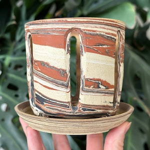 以下が含まれることがあります： 茶色、白、黒のまだら模様のセラミック製のプランター。プランターには受け皿と側面に3つの切り抜きがあります。