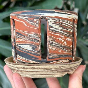 Puede incluir: Una maceta de cerámica de color terracota con un patrón de mármol y agujeros de drenaje. La maceta tiene un platillo a juego.