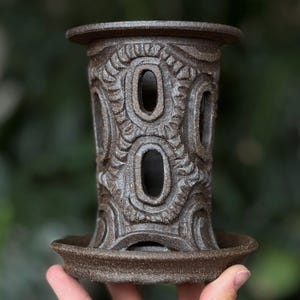 May include: A brown ceramic candle holder with an intricate carved design featuring oval cutouts. The candle holder has a cylindrical shape with a base and a top rim, and is approximately 15 cm tall.