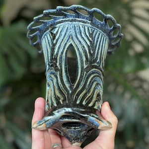 May include: A handcrafted ceramic vase with a unique design. The vase features a dark blue and black color scheme with intricate wave-like patterns and a decorative, openwork rim. The vase is held in a hand, showcasing its detailed craftsmanship.