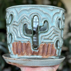 Puede incluir: Jardinera de cerámica hecha a mano con un esmalte azul claro. El diseño presenta formas de cactus recortadas y un patrón ondulado texturizado. La base tiene un platillo a juego. La jardinera tiene aproximadamente 15 cm de diámetro.