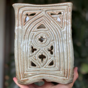 May include: A rectangular ceramic candle holder with a light gray glaze and a decorative, carved Gothic-style pattern. The design includes arched shapes, circles, and floral-like cutouts. The edges have a subtle brown hue.