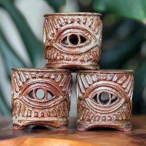 May include: Three stacked, handmade ceramic vessels with an eye design. The brown and cream-colored pottery has a textured surface and a carved eye in the center. The vessels are displayed on a wooden surface.