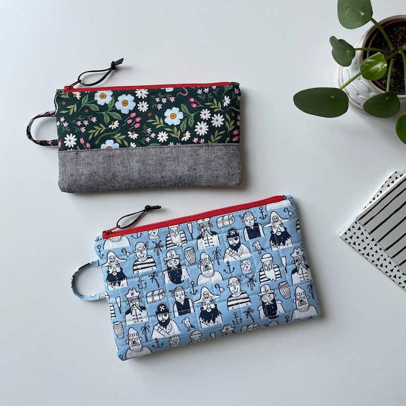May include: Two zippered pouches, one with a floral print in shades of green and white, and the other with a blue background and a repeating pattern of pirates and treasure chests.