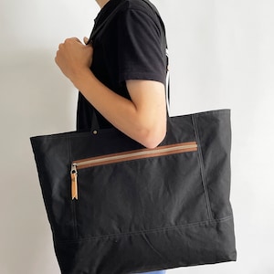 May include: A black canvas tote bag with a brown zippered pocket and leather accents. The bag has a top zipper closure and two long handles.