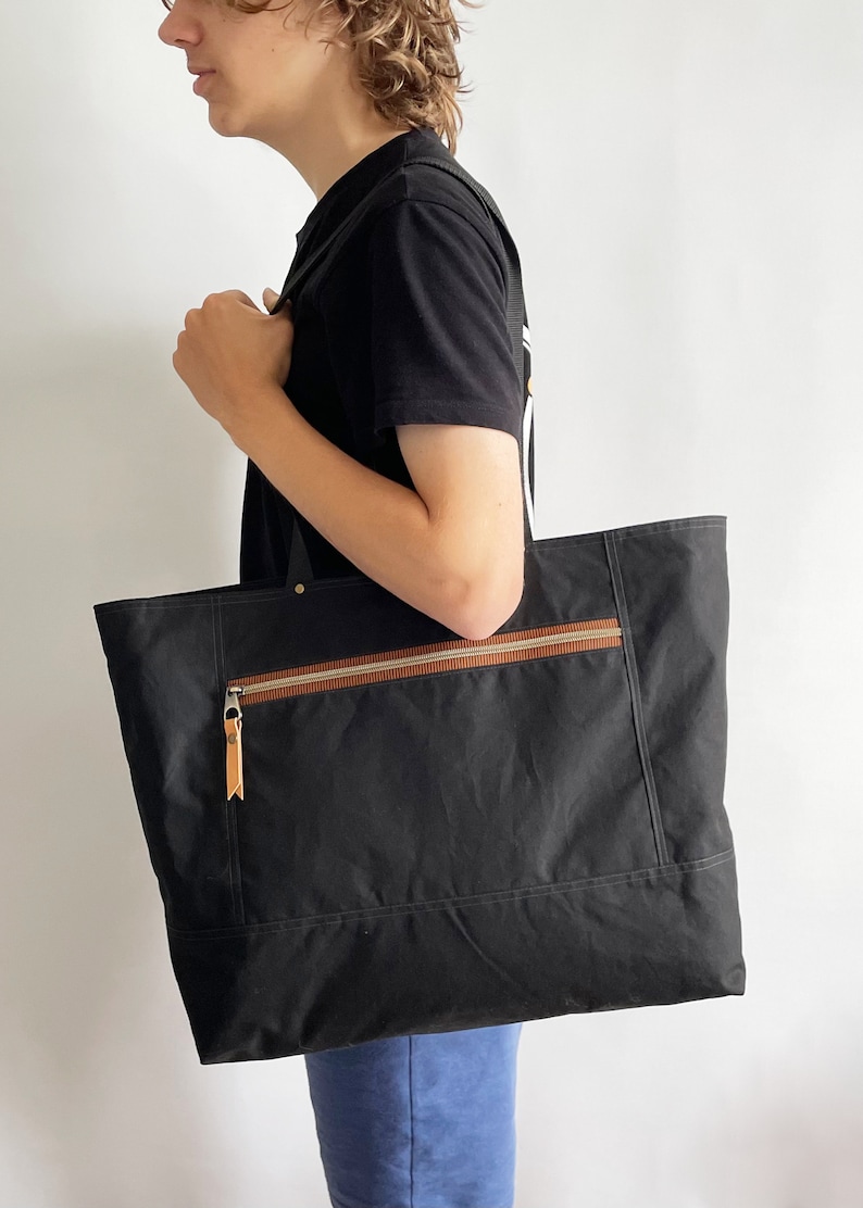 May include: A black canvas tote bag with a brown zippered pocket and leather accents. The bag has a top zipper closure and two long handles.