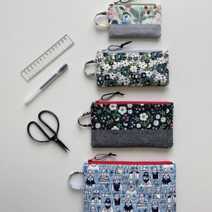 May include: Four zippered pouches with different fabric patterns. The pouches are lined with a gray fabric and have red zipper pulls. The pouches are arranged vertically from top to bottom. The top pouch has a floral pattern, the second pouch has a white flower pattern, the third pouch has a floral pattern with a red zipper pull, and the bottom pouch has a blue and white pattern with a red zipper pull.