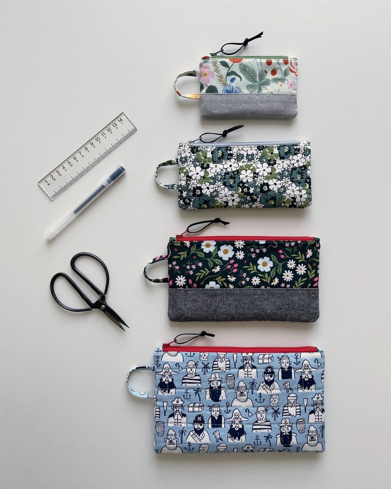 May include: Four zippered pouches with different fabric patterns. The pouches are lined with a gray fabric and have red zipper pulls. The pouches are arranged vertically from top to bottom. The top pouch has a floral pattern, the second pouch has a white flower pattern, the third pouch has a floral pattern with a red zipper pull, and the bottom pouch has a blue and white pattern with a red zipper pull.